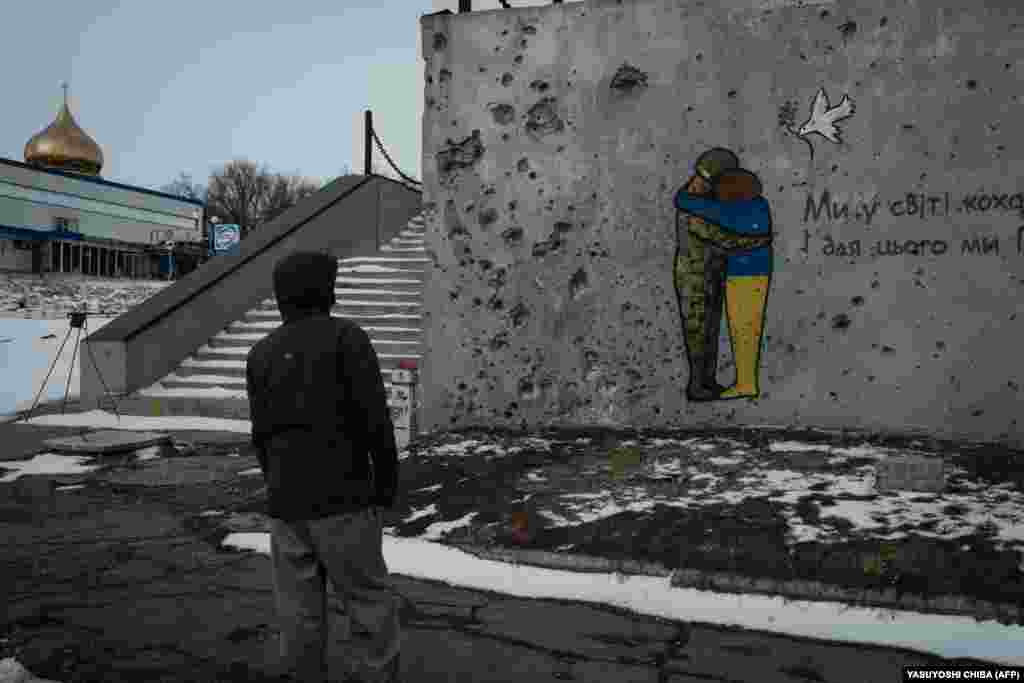 Чоловік дивиться на посічену осколками стіну, на якій намальований мурал із написом: «Ми у світі кохання помножимо. І для цього ми переможемо!». Куп'янськ, Харківська область, 13 лютого 2023 року