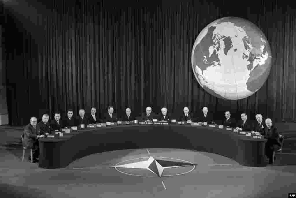 O conferință NATO la Paris în 1957, în spatele emblemei NATO, adoptată în 1953. Drapelul NATO are un albastru închis, reprezentând Oceanul Atlantic, cu o rozetă reprezentând direcția spre pace și un cerc simbolizând unitatea. 