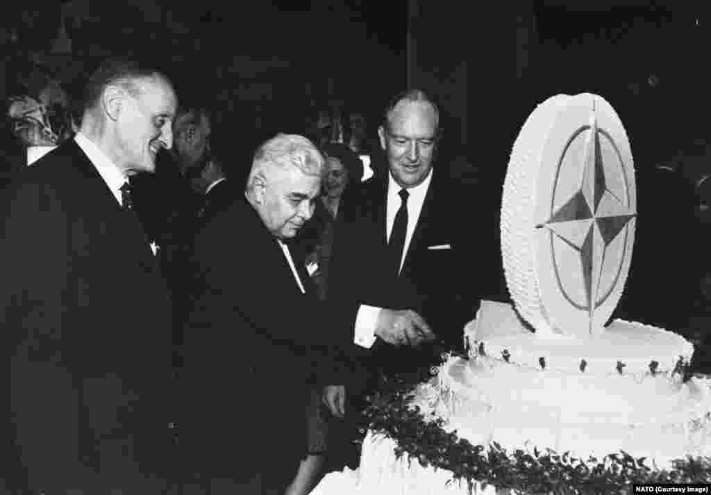 Secretarul general al NATO, Manlio Brosio (stânga), și secretarul de stat al SUA, William Rogers, îl privesc pe prim-ministrul islandez, Bjarni Benediktsson, cum taie un tort cu tematică NATO în timpul sărbătoririi celei de-a 20-a aniversări a alianței la Washington D.C. în 1969.